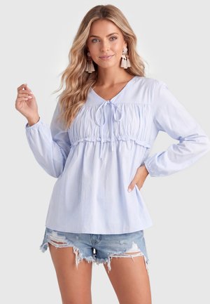Frau mit langen blonden Haaren, die eine hellblaue Bluse mit langen Ärmeln und Bindendetail sowie zerrissene Jeansshorts trägt und mit einer Hand in der Hüfte posiert.