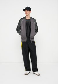 Sivi trening jakna s bijelim detaljima nosi se preko crne majice. Crne hlače imaju žute bočne pruge. Bijele tenisice upotpunjuju izgled.