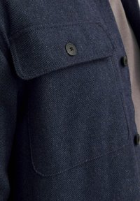 Giacca in tessuto testurizzato blu navy con tasca a pattina, dotata di chiusura con bottone nero e due ulteriori bottoni neri sul davanti.