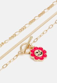 Colar em tom de ouro, apresentando três correntes: uma corrente grossa, uma corrente de cobra plana e uma corrente delicada com um pendente de flor sorridente rosa.