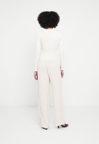 Femme aux cheveux bouclés portant un haut blanc à manches longues, un pantalon beige large et des chaussures marron à talons, debout face à un mur blanc.