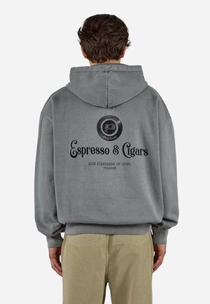 Mand iført grå hættetrøje med "Espresso & Cigars" tekst og logo på ryggen, kombineret med beige bukser, vendt væk.