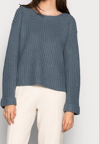 Kvinna iförd en blå grovstickad tröja med uppvikta ärmar och ljusbeiga byxor mot en enhetlig bakgrund.