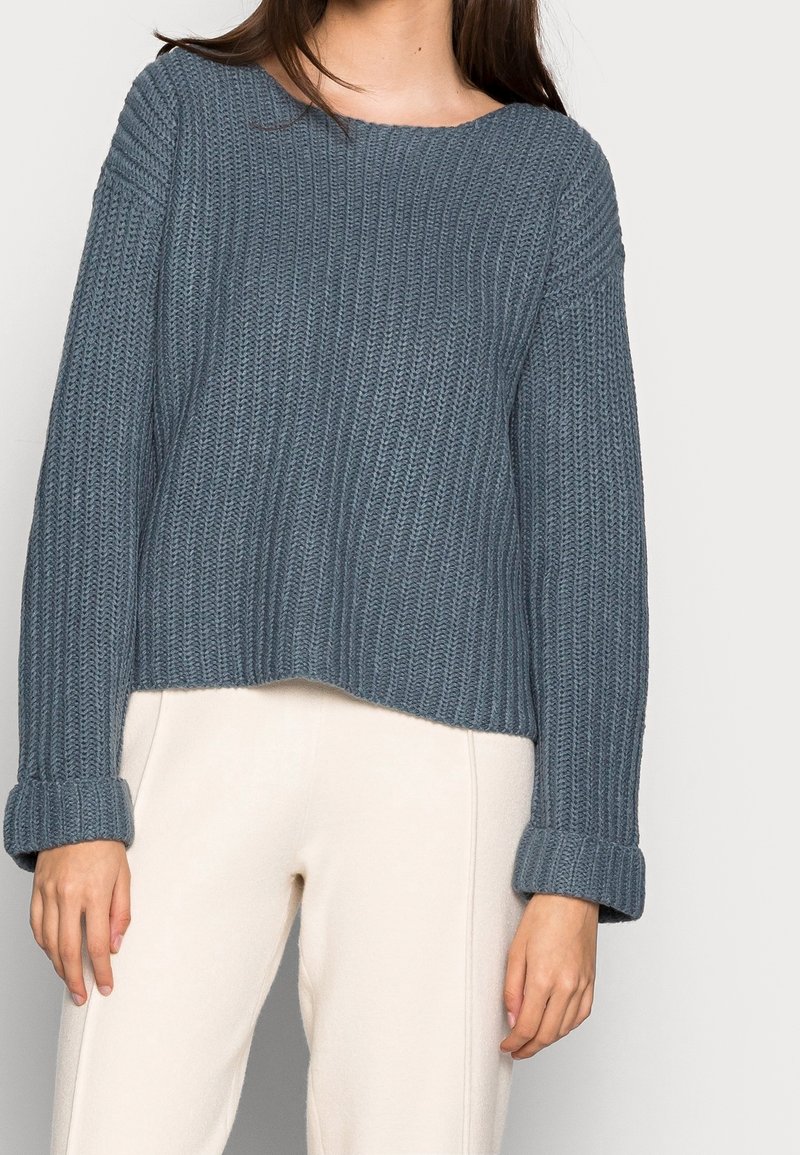 Kvinna iförd en blå grovstickad tröja med uppvikta ärmar och ljusbeiga byxor mot en enhetlig bakgrund.