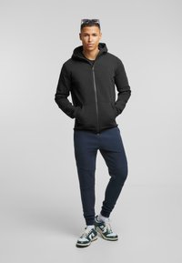 Felpa nera con zip e tasche frontali, abbinata a pantaloni jogger blu e sneakers verde e bianche. Il modello si trova in piedi con le mani nelle tasche.