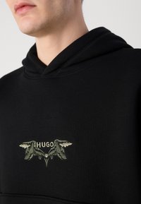 Schwarzer Kapuzenpullover mit einem zentralen, gestickten Design mit 'HUGO' und geflügelten Akzenten in sanftem Grün und Creme. Weicher, strukturierter Stoff.
