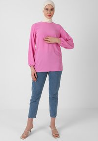 Top rosa a maniche lunghe con polsini elasticizzati, abbinato a jeans azzurri. Il modello indossa un hijab beige e sandali open-toe. Design semplice.