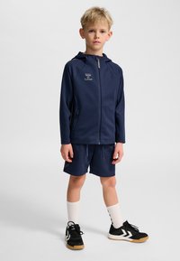 Mörkblå sportig hoodie med hel dragkedja och sidofickor, tillsammans med matchande shorts. Svarta träningsskor med vita detaljer och crew-strumpor.