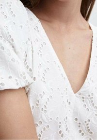 Blouse blanche à manches courtes bouffantes, comportant des motifs floraux découpés complexes et un col en V. Texture douce, tissu léger.