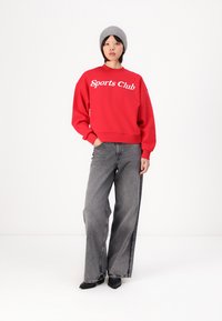 Felpa rossa con la scritta "Sports Club" in bianco, jeans grigi a gamba larga e scarpe nere. Un berretto grigio completa il look casual.