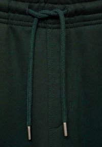 Zelene bombažne nogavice imajo vrvico z kovinskimi konicami, elastičen pas in teksturirano tkanino, zasnovano za udobje in fleksibilnost.