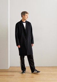 Man wearing a black long coat, black track pants with white stripes, and black sneakers standing on a wooden floor against a plain wall.