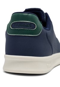 Blå lädersneaker med grön hälflik, vit sula och perforerad sidodetalj. Logotypen är präglad på hälen.