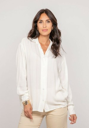 Femme aux longs cheveux foncés portant une chemise blanche ample à boutons et un pantalon beige, debout devant un fond clair uni.