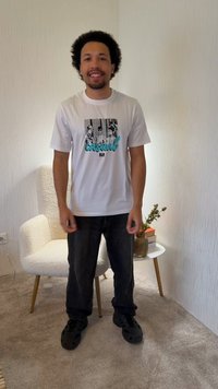 Homme aux cheveux bouclés portant un t-shirt blanc à motif graphique et un pantalon noir, debout en souriant dans une pièce avec un fauteuil blanc et une petite table avec un vase et des livres.
