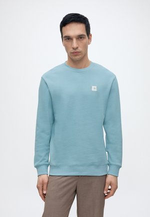 Young man wearing light blue crewneck sweatshirt and brown pants, standing against plain light background, facing forward with neutral expression.