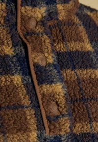 Brown, blue, and yellow checkered fleece fabric with a soft texture; features plastic snap buttons and a contrasting brown trim along the edge.