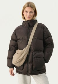 Daunenjacke in dunkelbraun mit hohem Kragen, Vorderreißverschluss, Seitentaschen und elastischen Bündchen. Kombiniert mit einer beigen Umhängetasche.
