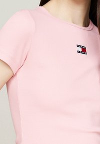 Pink ribbed short-sleeve shirt with a round neckline, featuring a small embroidered "Tommy Jeans" patch in navy, white, and red.