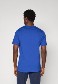 Blå bomulds T-shirt med rund hals og korte ærmer, der har en glat tekstur og en lige kant, set fra ryggen.