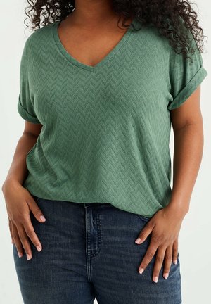 Kobieta ubrana w zielony, teksturowany top z krótkim rękawem i dekoltem w serek oraz ciemnoniebieskie dżinsy, z rękami spoczywającymi blisko kieszeni.
