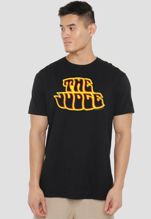 Schwarzes Baumwoll-T-Shirt mit einem großen, auffälligen, gelb-orangefarbenen Grafiktext "DER JUROR" auf der Vorderseite. Mit kurzen Ärmeln und Rundhalsausschnitt.