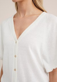 Cardigan bianco leggero con scollatura a V e maniche corte. Caratterizzato da una chiusura con bottoni e un sottile punto di maglia testurizzato.