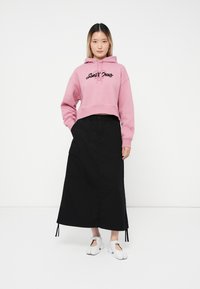 Felpa rosa cropped con logo nero "Santa Cruz", abbinata a una lunga gonna nera con tasche cargo e scarpe bianche slip-on.