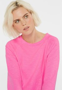 Blonde Frau mit kurzen Haaren, die ein leuchtend pinkes Langarmshirt trägt und leicht zur Seite schaut, vor einem weißen Hintergrund.