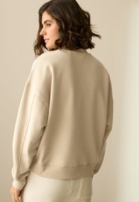 Bež sweatshirt opuštenog kroja, spuštenih ramena i rebrastih manžeta. Glatka tekstura, jednobojan, bez vidljivih uzoraka ili naglasaka.