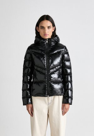 Schwarze glänzende Daunenjacke mit Kapuze, mit gesteppten Abschnitten und einem Reißverschluss vorne. Getragen über cremefarbenden Hosen.