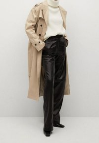 Beżowy trencz z podwójnymi guzikami, noszony na białym swetrze z prążkami i czarnych, błyszczących szerokich spodniach z butami na wysokim obcasie w kształcie szpica.