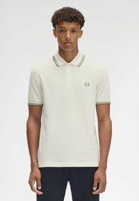 Camisa polo de algodón color blanco roto con detalles en el cuello y puños a rayas verdes, que presenta un sutil logo bordado en el lado izquierdo del pecho.