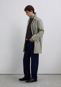 Homme se tenant à l'intérieur portant un trench beige, un pull noir, un jean bleu marine, une ceinture noire et des chaussures en cuir noires, avec une main dans la poche.