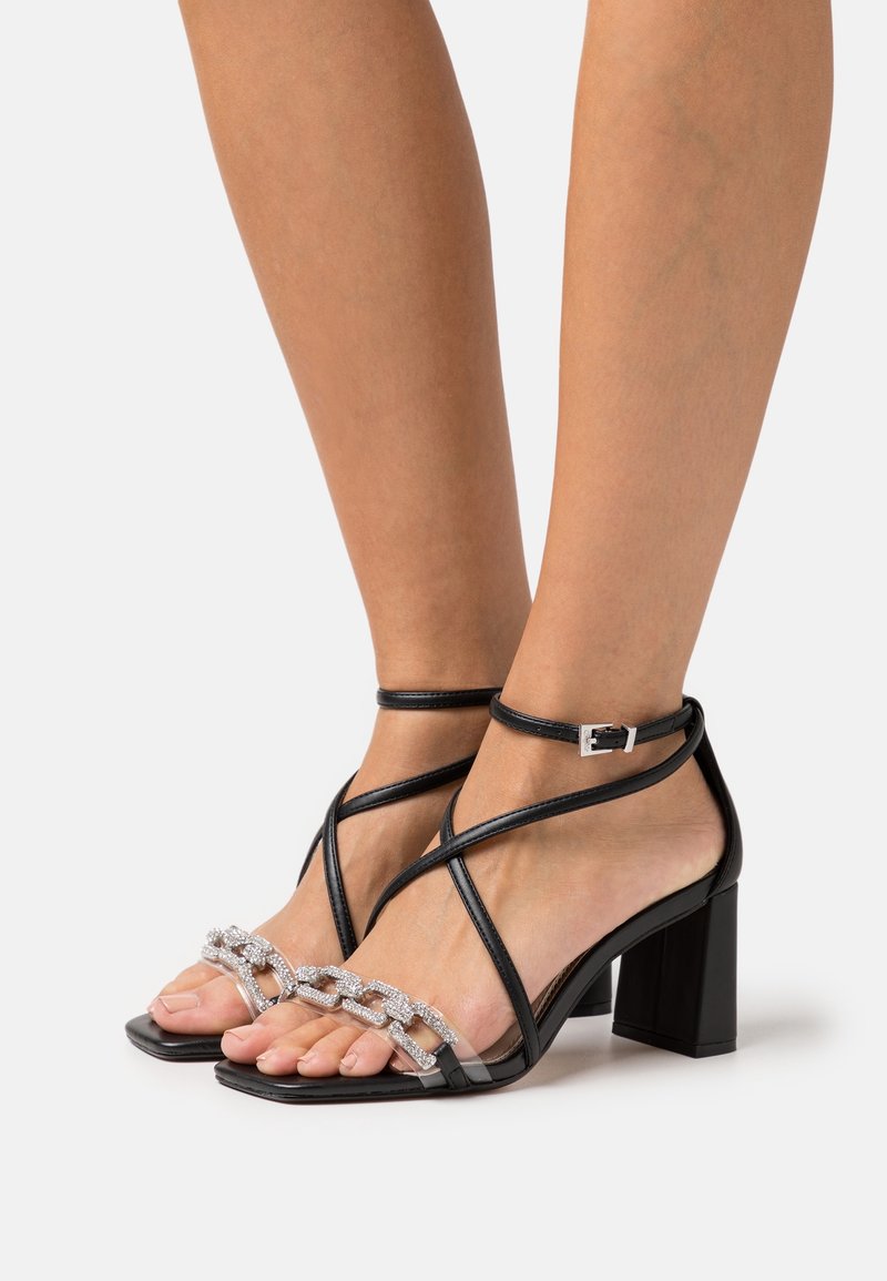 Sandales noires à talons hauts avec brides à la cheville et détail chaîne de strass sur les orteils, portées pieds nus sur fond blanc.