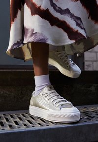 Stříbrné metalické tenisky s hladkým povrchem, bílou gumovou podrážkou a třemi pruhy. Nošené s bílými ponožkami a vzorovanou sukní.