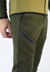 Pantalones deportivos en verde oliva con un diseño ajustado, que cuentan con un bolsillo con cremallera y un acento de logo negro. La tela parece texturizada y suave.
