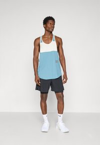 T-shirt sem mangas azul claro e branca com uma bainha arredondada, combinada com calções pretos. A parte de cima apresenta uma pequena etiqueta de logotipo perto da bainha.