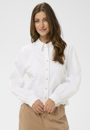 Chemise blanche boutonnée avec manches longues, ornée d'une bordure en dentelle florale à l'ourlet et de boutons couleur beige, portée avec un pantalon beige.