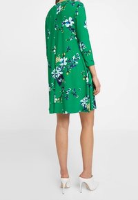 Green floral dress with three-quarter sleeves, featuring a floral pattern in white and blue. Cut above the knee, finished with a pleat in the back.