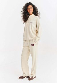Béžová mikina s polovičním zipem a vyšívaným logem, doplněná odpovídajícími béžovými kalhotami s logovou nášivkou. Model má na sobě hnědé tenisky.