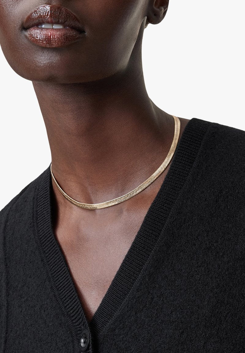 Gul choker halskæde med en glat, skinnende tekstur, båret med en sort, struktureret cardigan med V-udskæring og knaplukning.