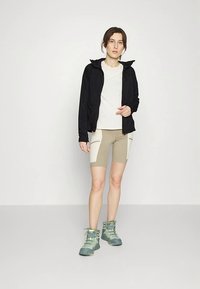 Pārgājiena apģērbs ar melnu vieglu jaku, baltu T-kreklu, bēšām šortām ar krēma paneļiem un tirkīza pārgājienu zābakiem ar auklām.