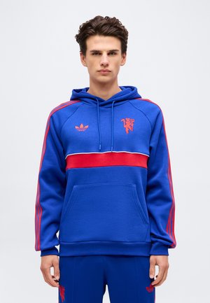 Jeune homme portant un sweat à capuche bleu avec des rayures rouges, un logo rouge et une poche devant, debout devant un fond clair uni.