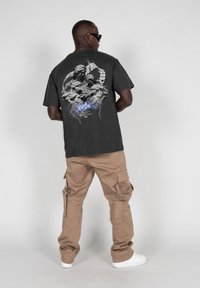 Mørkegrå grafisk T-shirt med et detaljeret design på ryggen, parret med beige cargobukser og hvide sneakers. Glat stoftekstur.