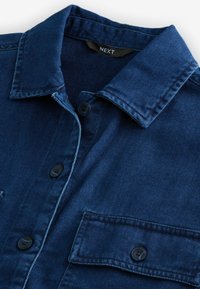 Niebieska koszula dżinsowa z ostrym kołnierzykiem, czarnymi guzikami i kieszenią na piersi z widocznym ściegiem. Miękka tekstura i tkanina splotowa.