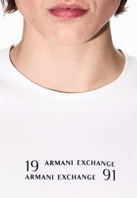 Weißes T-Shirt mit rundem Ausschnitt. Mit schwarzem Text: "19 ARMANI EXCHANGE" und "ARMANI EXCHANGE 91." Glatte Textur, lässiges Design.