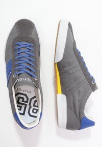 Baskets en toile grise avec accents bleus, semelle en caoutchouc blanche, lacets bleus et détail jaune sur le talon. Le design à panneaux cousus offre une texture.