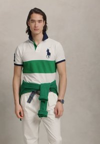 Baltā polo krekls ar zaļām horizontālām svītrām, tumši zils apkakle un zils zirga logo. Zaļš adījuma džemperis sasiets ap vidukli. Baltas bikses.