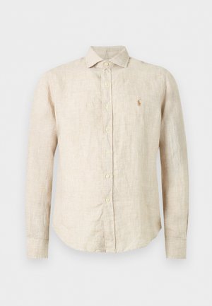 Jeune homme debout, portant une chemise en lin beige, un pantalon blanc ample et des chaussures beiges, les mains partiellement dans les poches, fond neutre.